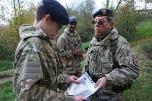 An Adult Volunteer talks to a young Cadet holding a map. An older Cadet is stood in the background.
