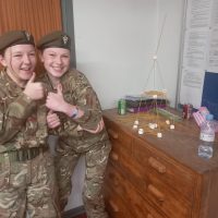 Two female Cadets in uniform with thumbs up, beside a tower made of spaghetti and marshamallows.