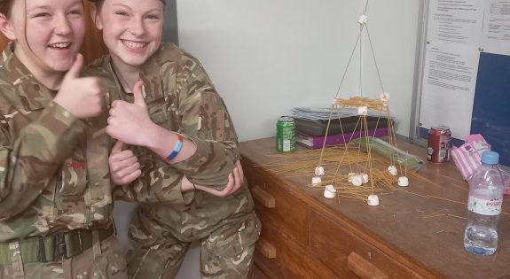 Two female Cadets in uniform with thumbs up, beside a tower made of spaghetti and marshamallows.